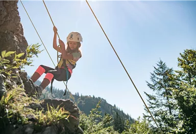 Ein Kind klettert an einer Felswand und nutzt ein Seil zur Sicherung. Die Umgebung ist von Bäumen und Bergen umgeben, und der Himmel ist klar und sonnig.