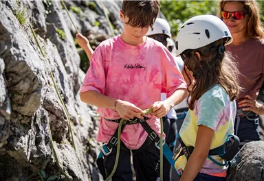 Kinder bereiten sich auf das Klettern vor, während sie Helme tragen. Sie stehen an einer Felswand und sind mit Sicherheitsausrüstung ausgestattet.