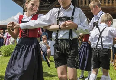 Ein fröhlicher Tanz von Kindern in traditioneller bayerischer Tracht. Im Hintergrund steht ein Holzhaus und die Sonne scheint.