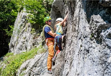 Ein Kletterer hilft einem Kind beim Klettern an einer Felswand. Die Sonne scheint, und die Umgebung ist grün und naturnah.