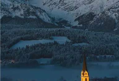 An idyllic mountain landscape with snow-capped peaks and a small village. The church in the village glows softly in the dark.