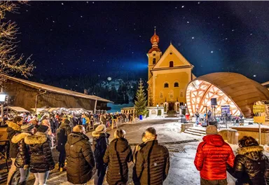 A festive Christmas market with many visitors under the starry sky. In the background stands a church and a stage presenting music.