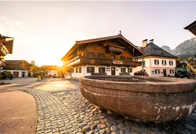 Eine malerische Landschaft mit traditionellen Holzhäusern und einem alten Brunnen. Die Sonne geht hinter den Bergen auf und schafft eine warme Atmosphäre.