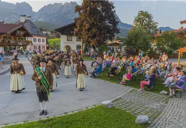 A traditional festival with a brass band and dancing people in traditional costumes. In the background, an enthusiastic audience sits in chairs.