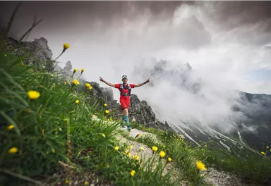 Ein Sportler in roter Kleidung steht auf einem Wanderweg in den Bergen. Umgeben von grünen Wiesen und Wolken strahlt er Freude aus.