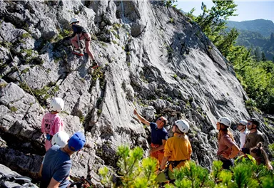 Eine Gruppe von Menschen beobachtet einen Kletterer an einer Felswand. Der Hintergrund zeigt grüne Bäume und Berge im Freien.