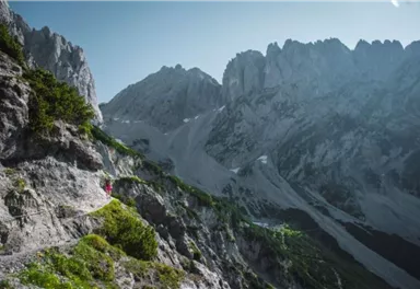 An impressive mountain landscape with tall cliffs and snow-covered peaks. A hiker follows a narrow path between the mountains.
