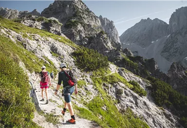 Two hikers are walking a steep path in the mountains. The landscape is impressive with tall rocks and green meadows.