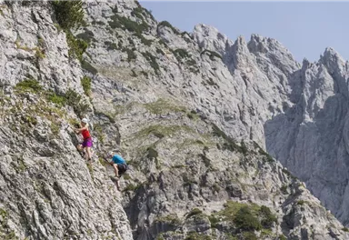 Zwei Kletterer erklimmen eine steile Felswand in einer beeindruckenden Berglandschaft. Der Himmel ist klar und die Umgebung wirkt majestätisch.