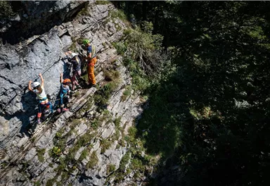 Eine Gruppe von Kletterern bewegt sich an einer steilen Felswand. Die Umgebung ist grün und bewaldet, was die natürliche Schönheit der Szene betont.