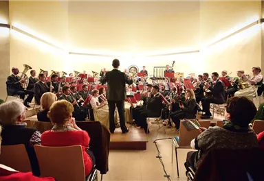 A large orchestra is playing on a stage in front of an audience. The conductor is directing the ensemble in a festive concert hall.