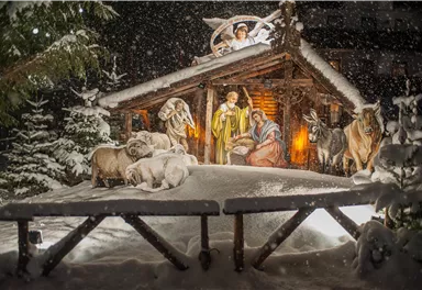 Eine festliche Krippe im Schnee mit Figuren von Maria, Josef und dem Jesuskind. Rundherum sind Tiere und schneebedeckte Bäume zu sehen.