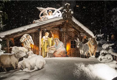 Eine winterliche Krippe mit Maria, Josef und dem neugeborenen Jesus in einem Stall, umgeben von Tieren. Sanfte Schneeflocken fallen auf die Szene und erzeugen eine festliche Atmosphäre.
