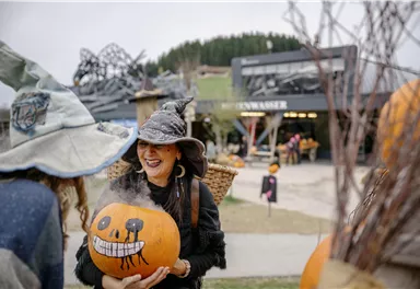 A woman with a witch's hat smiles and holds a pumpkin with a face. In the background, other visitors and a decorated environment can be seen.