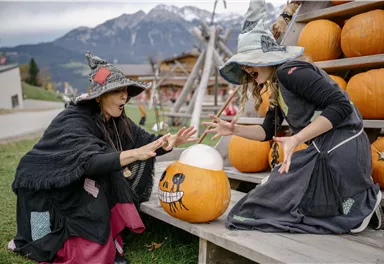 Two women in witch costumes are standing in front of a wooden shelf with pumpkins. They seem to be looking at a pumpkin with a face and appear to be excited.