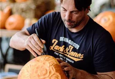 Ein Mann schnitzt mit einem Werkzeug in einen Kürbis. Im Hintergrund sind weitere Kürbisse und Strohballen zu sehen.