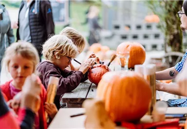 Zwei Kinder schnitzen mit Begeisterung Kürbisse an einem Tisch. Im Hintergrund sind weitere Kinder und Erwachsene zu sehen, die ebenfalls an der Kürbisaktion teilnehmen.