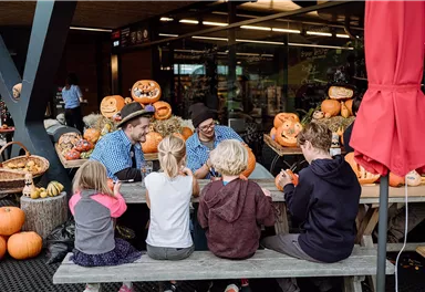 Eine Gruppe von Kindern sitzt an einem Tisch und schaut zu, während Erwachsene an Kürbissen arbeiten. Im Hintergrund sind viele dekorierte Kürbisse zu sehen.