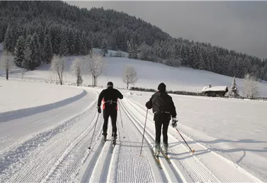 Two skiers in winter clothing glide on a freshly prepared trail through a snow-covered landscape. In the background, there are trees and a small cabin.