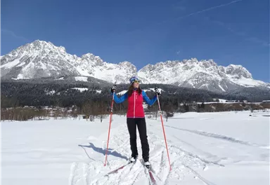 A woman is standing in the snow and cross-country skiing with ski poles. In the background, impressive mountains and a blue sky can be seen.