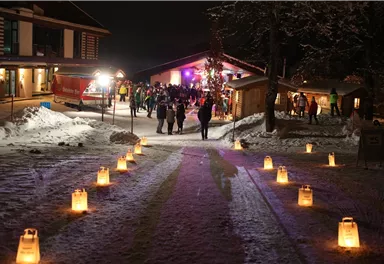 Eine festlich illuminierte Winterlandschaft mit schneebedeckten Wegen. Gruppen von Menschen versammeln sich um eine gemütliche Hütte unter dem nächtlichen Himmel.