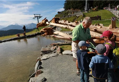 An older man is explaining something to children at the shore of a small body of water in the mountains. In the background, a mountain railway and a beautiful landscape with mountains can be seen.