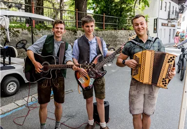 Drei Männer in traditioneller Kleidung spielen Musikinstrumente auf einer Straße. Sie haben einen Akkordeon, eine Gitarre und eine E-Bass dabei.