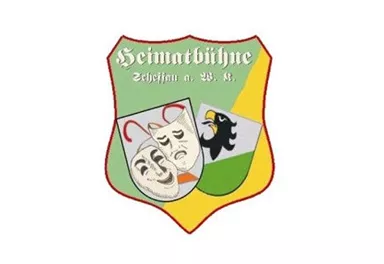 Ein Wappen mit dem Schriftzug "Heimatbühne" und verschiedenen Symbolen, darunter ein Theatermaskenmotiv und ein schwarzer Vogel. Die Farben sind leuchtend und in einem Schild-Design angeordnet.