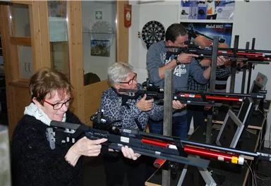 Eine Gruppe von Personen zielt mit Sportgewehren in einem Trainingsraum. Die Atmosphäre ist konzentriert und sportlich.