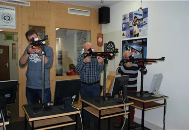 Drei Männer stehen an Schießständen und zielen mit Luftgewehren. Im Hintergrund sind Computer und eine Pinwand sichtbar.