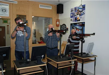 Drei Männer stehen an Schießständen mit Gewehren. Sie konzentrieren sich auf das Zielen und Schießen, während Computer vor ihnen stehen.