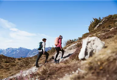 Zwei Wanderer erklimmen einen felsigen Weg in den Bergen. Die Landschaft ist schön und der Himmel ist klar und blau.