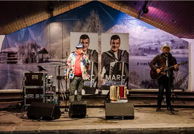 Ein Live-Auftritt zweier Musiker auf einer Bühne. Im Hintergrund sind große Plakate von Marc Pichler zu sehen.