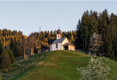 A small church on a hill, surrounded by trees. The landscape is green and the atmosphere is tranquil.