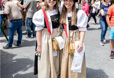 Zwei Mädchen in traditionellen Trachten mit schwarzen Hüten und goldenen Röcken. Sie lächeln fröhlich vor einer Menschenmenge auf einem Fest.