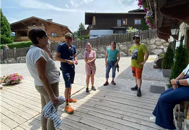 Eine Gruppe von Menschen steht auf einem Holzsteg und unterhält sich. Im Hintergrund sind traditionelle Gebäude und Blumenpflanzen zu sehen.