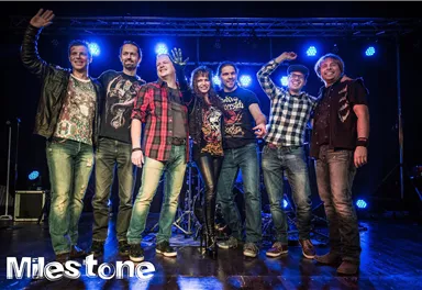 Eine Gruppe von sieben Musikern steht auf einer Bühne. Sie lächeln und posieren vor einem festlichen Hintergrund mit blauen Lichtern.