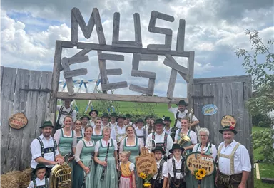 Eine Gruppe von Menschen in traditionellen Trachten steht vor einem festlichen Eingangsschild mit der Aufschrift "MUSIKFEST". Sie lächeln und posieren für das Foto in einer ländlichen Umgebung.