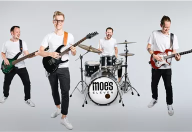 Eine Band mit vier Mitgliedern steht in einem hellen Raum. Sie spielen verschiedene Instrumente und lächeln in die Kamera.