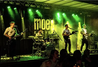 Eine Band spielt Live-Musik auf einer Bühne mit grünen Lichtern. Im Hintergrund ist ein großes Schild mit dem Namen "moe's" zu sehen.