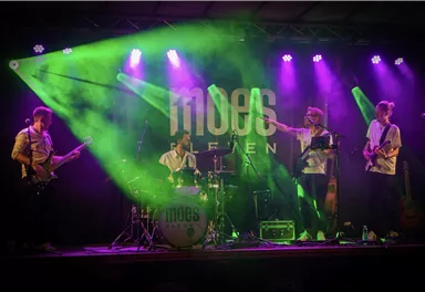 Eine Band spielt auf einer Bühne mit bunten Lichtern. Die Musiker sind enthusiastisch und die Atmosphäre ist lebhaft.