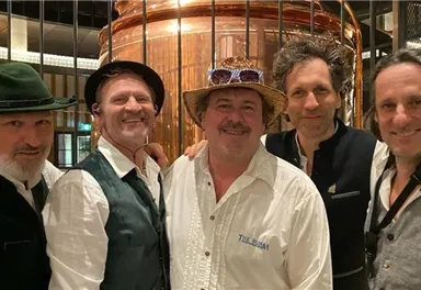 Eine Gruppe von fünf Männern steht lächelnd vor einer großen Kupferstruktur. Alle tragen festliche Kleidung und scheinen eine gute Zeit zu haben.