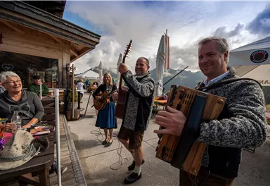 Eine Gruppe von Musikern spielt traditionelle Musik vor einer Berghütte. Im Hintergrund sind tanzende Menschen und eine malerische Berglandschaft zu sehen.