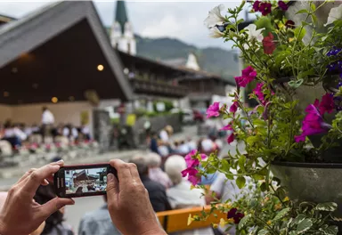 Eine Person hält ein Handy und fotografiert eine Veranstaltung im Freien. Im Vordergrund sind bunte Blumen zu sehen, während im Hintergrund die Menschenmenge und eine Berglandschaft zu erkennen sind.