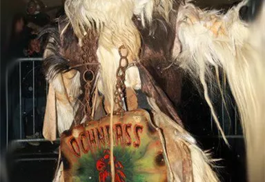 A spooky, masked character with long, shaggy fur and a threatening expression. He holds a sign that says "Oachkatzlschwoaf".