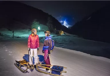 Zwei Personen stehen auf einem verschneiten Weg bei Nacht, bereit zum Schlittenfahren. Im Hintergrund sind schneebedeckte Berge und Lichter eines Skigebiets sichtbar.