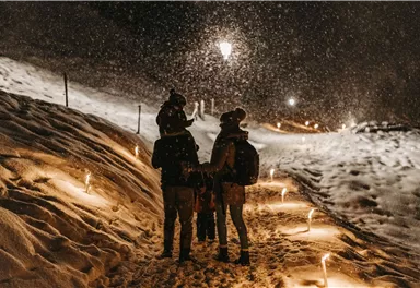 A family strolls through the snow at night as snowflakes fall. The path is lined with lights that create a festive atmosphere.