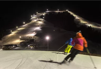 Eine nächtliche Skisession auf einer beleuchteten Piste. Skifahrer fahren den Hang hinunter, während die Lichter die Landschaft erhellen.