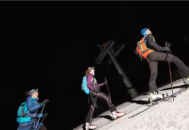 Three skiers are climbing the slope at night. They are wearing ski and safety gear, while a ski lift is visible in the background.