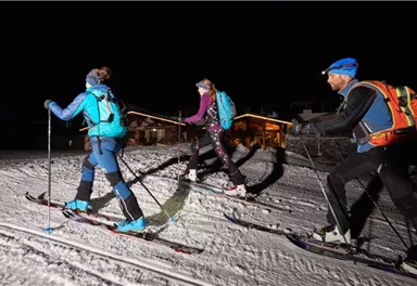 Three people are skiing with touring skis in the snow at night. In the background, illuminated buildings can be seen.